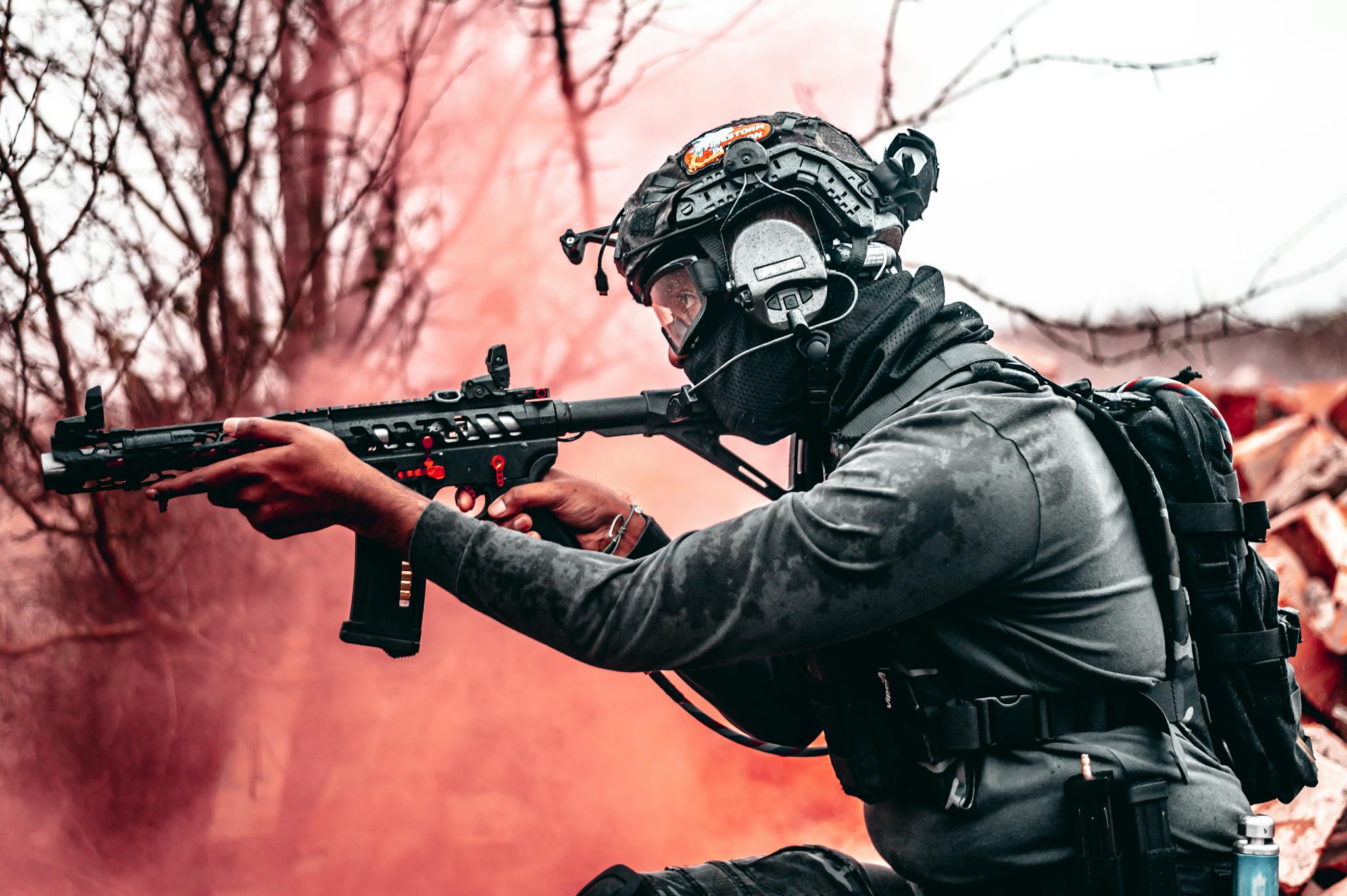 A tactical soldier in full combat gear aims a rifle amidst smoke outdoors.